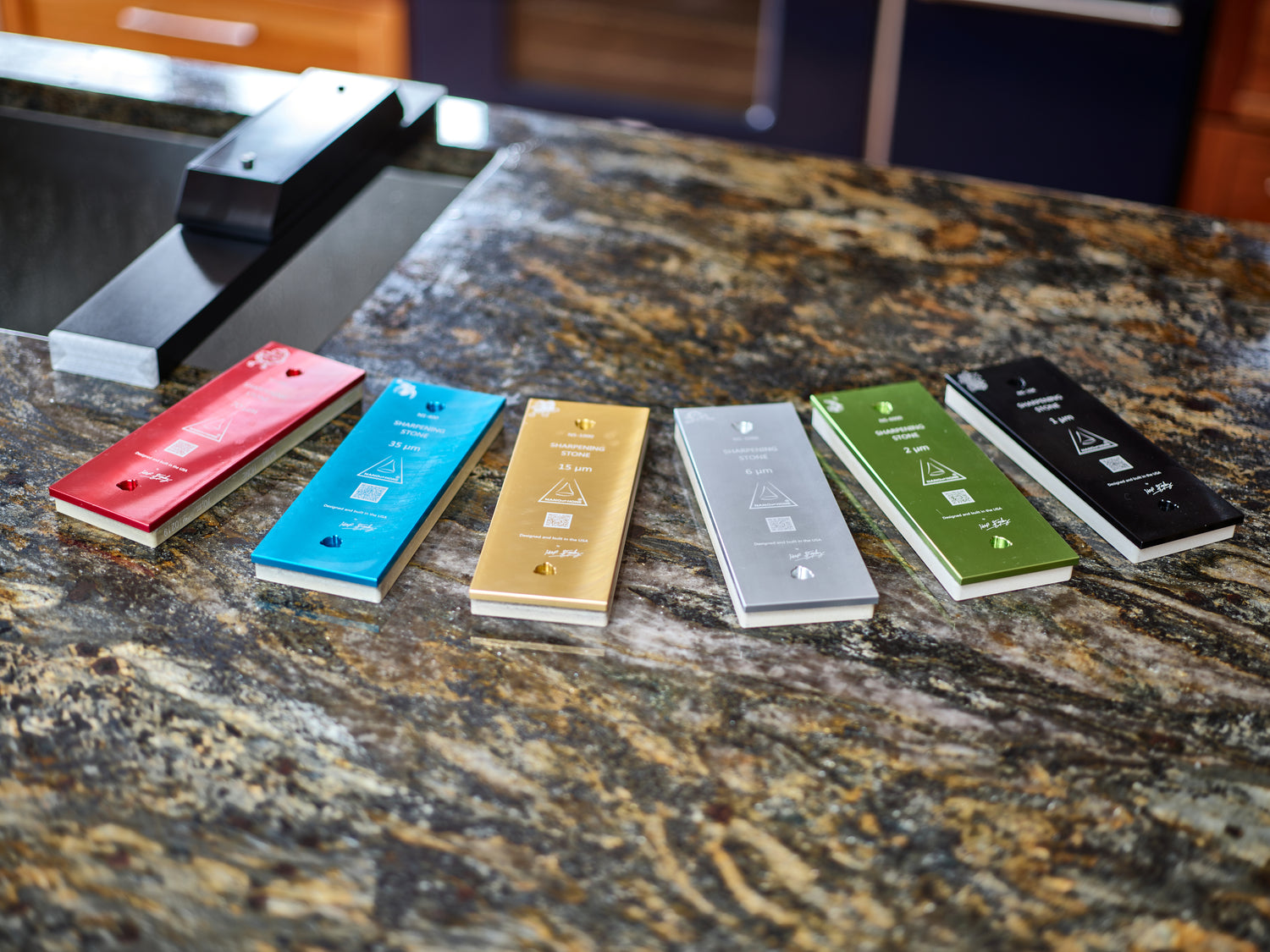 Sharpening stones lay flat on a granite countertop with a Sink Bridge sharpening stone holder over a sink in the background