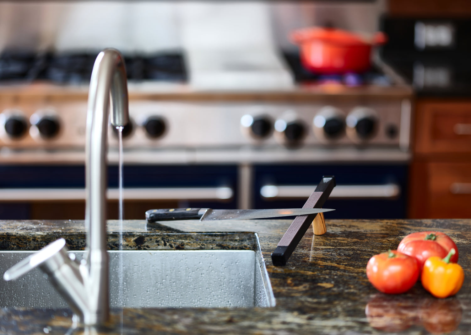 A sharp knife rests on a diamond ceramic kitchen strop in a kitchen with trickling water from a sink and tomatoes in the foreground